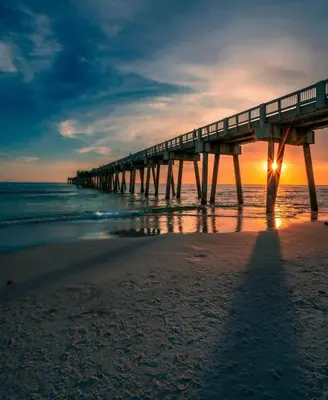 a pier on the beach with the sun setting at Dolphin View at 8629 Surf Drive by Nautical Properties in Panama City Beach