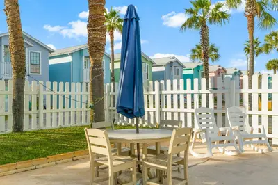 a table with a blue umbrella and chairs and a fence at Sand Castle West #G6 in Panama City Beach