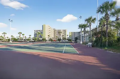 a tennis court with palm trees and buildings in the background at Oceanfront Condo - Balcony Views & Beach Access in Panama City Beach